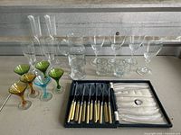 Group view of colored cordial stems, clear wine glasses, champagne flutes, pitcher, small mugs, and boxed Sheffield cutlery set