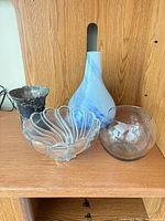 Four items on shelf: metal vase, clear swirl glass bowl, blue-white art glass vase, clear fishbowl vase