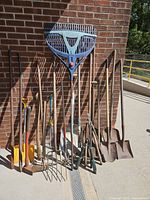 Full group of long-handled garden tools leaning against brick wall