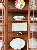 Cabinet shelf showing floral round plate, blue floral oval bowl, scalloped white rectangular platter, pink floral oval platter