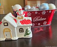 Front view of ceramic Santa house beside red metal Believe basket filled with ornaments