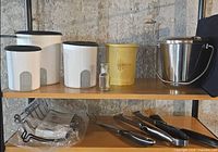 Shelf with three white canisters, yellow canister, glass spice jar, stainless ice bucket, roasting rack in plastic, BBQ tools