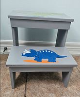 Front view of two-tier grey wooden step stool showing blue triceratops painting on lower tread