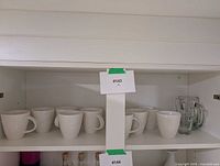 Shelf view showing groups of white ceramic mugs and two clear glass mugs