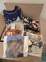 Box containing folded graphic tees showing various designs