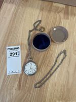 Group shot showing Alpina pocket watch attached to one chain, separate second chain, storage container with lid, and lot tag