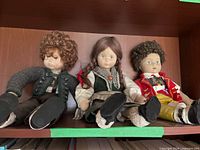 Three cloth dolls seated on shelf displaying full outfits and facial details