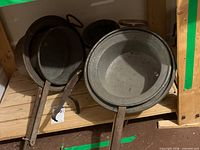 Two large and one smaller metal pour pots with long hooked handles resting on wooden shelf