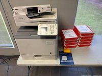 Printer with toner box atop and stack of copy paper plus manual on table