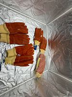 Four pairs orange suede gloves with yellow cuffs displayed, one pair sealed