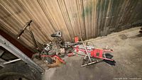 Pile of disassembled mini bike parts including engines, frame, fork, wheels and seat against wall