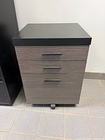 Front view showing three drawers, black top, gray wood-grain fronts, wheels