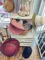 Shelf with multiple hats including straw sun hat, pink straw hat, felt hats, burgundy fedora, and black riding helmet
