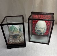 Two eggshell artworks each in its own glass wood case