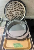 Stack showing perforated pizza pan, plain pizza pan, round cake pan, grey rectangular pan and gold sheet pan