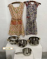 Full lot showing two aprons hanging and four stainless cookware pieces on pedestal