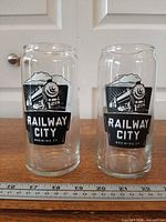 Two identical clear can-shaped beer glasses with Railway City Brewing logo beside ruler for scale