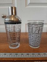 Shaker with metal top beside printed mixing glass, ruler for scale
