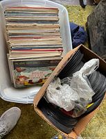 Plastic tote filled with vertically stacked LP records and adjacent cardboard box of loose 45s