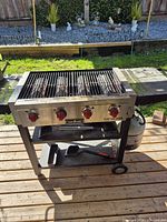 Front view of Camp Chef grill showing four red knobs, side shelf, propane tank, grill brush on bottom shelf