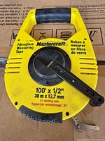 Front view of yellow Mastercraft 100 ft fibreglass tape measure showing brand, length, width, winding arm