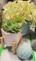 Group shot of faux succulents in pink planter on wooden stand, hydrangea arrangement in background, turquoise ribbed orb in foreground