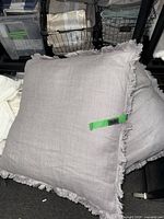 Two large grey square pillows with fringe on floor