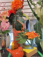 Group shot showing tall orange vase with florals, clear glass vase with calla lilies, orange bird, small orange glass vase, yellow box