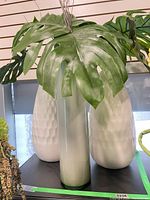 Front view showing grey cylinder vase flanked by two white textured vases, all with faux monstera leaves