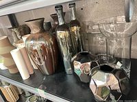 Shelf with assorted vases: glazed ceramic, metallic glass bottles, faceted silver ceramics, clear glass pieces, pastel cylinders