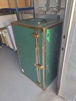 Front perspective of green steamer trunk standing upright, showing brass-tone edges, latches and side handle