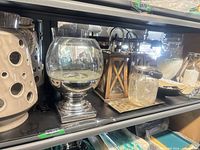 Shelf view showing ceramic lanterns, glass hurricane on silver base, wooden lanterns, seeded glass jar, metallic tray