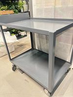 Overall view of grey metal two-tier trolley showing top surface, lower shelf and wheels