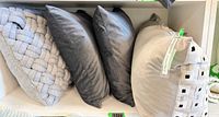 Four cushions on shelf: braided light grey, two dark grey, beige rectangle