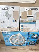 Stacked boxes: two Black+Decker pedestal fans, one Lasko Wind Machine 3300, two Lasko Breeze Machine
