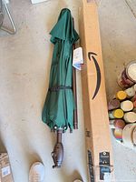 Folded green patio umbrella beside Amazon shipping box, showing crank handle, tilt joint and canopy