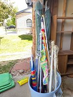 Blue bin containing poles, umbrellas, flags and umbrella base with green covers and yellow sand bag at side