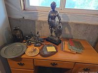 Desk top showing large knight statue, small figurines, platter, lantern, plaques, books