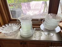 Group shot of all clear pressed glass pieces on counter showing platter, plates, trays, bowls, cup and cake stand