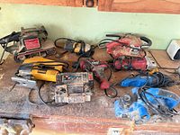 Group of assorted corded tools on bench including belt sander, jigsaw, charger, planer, drills and saw