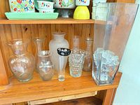 Eight assorted clear and frosted glass vases on wooden shelf, including tall rectangular column and various shapes