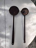 Two iron blacksmith ladles side by side on white surface