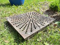 Top view of weathered square metal grate lying on grass showing radial hole pattern