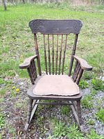 Front view of wooden pressed back rocking chair showing carving, spindles, arms, seat cushion, and rocker base