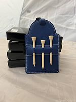 Front view of one blue leatherette golf tag with three wooden tees, black boxes stacked behind