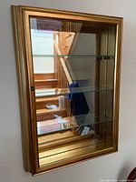 Front view of gold gilded wall-mounted curio cabinet showing glass door and three shelves.