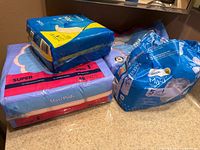 Assorted maxi pad packs on countertop, showing two purple 48-count packs, one blue/yellow 24-count pack, and opened blue bag