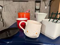 group view of saucer, daisy mug, red cup, orange planter, vase, square planter