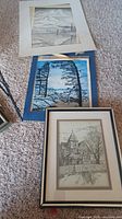 Group view showing all three artworks: framed house drawing, blue-mat lake scene, white-mat mountain scene