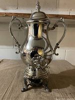 Full view of silver-plated coffee urn showing handles, spigot, lid, and ornate stand
